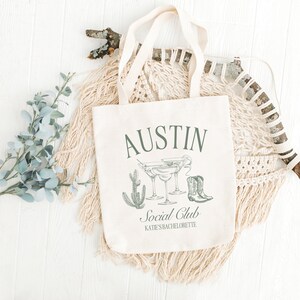 May include: A white canvas tote bag with the text "Austin Social Club Kate's Bachelorette" printed on it. The bag features a green illustration of cacti, margarita glasses, and cowboy boots.