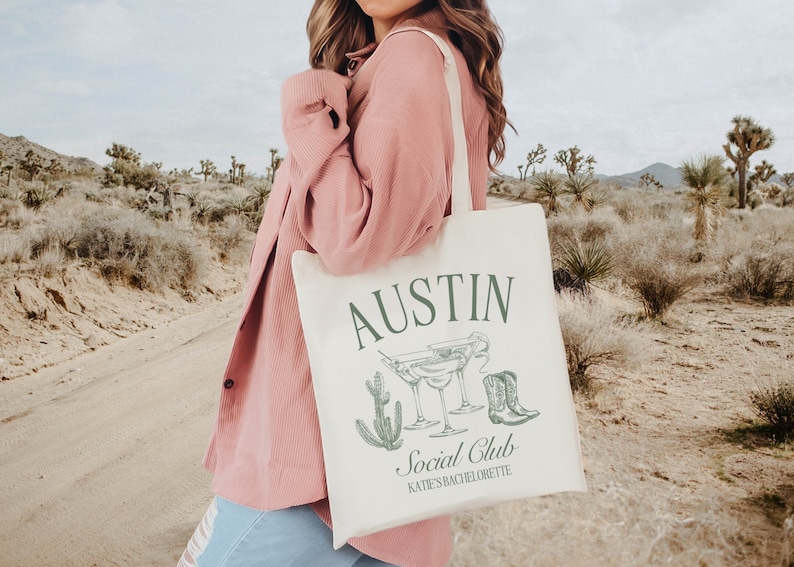 May include: A white canvas tote bag with the text "Austin Social Club" and "Katie's Bachelorette" in green lettering. The bag also features a cactus, margarita glasses, and cowboy boots.