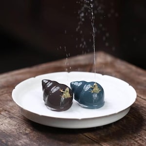 May include: Two ceramic frog figurines, one brown and one blue, sit on a white scalloped dish. A stream of water flows over the figurines. The frogs are small and detailed, adding a whimsical touch to the scene.