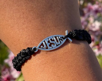May include: A black braided bracelet with a silver Jesus fish charm. The bracelet is worn on a person's wrist.