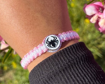 May include: A pink and white braided bracelet with a silver charm featuring a black spider design.