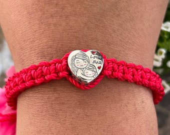 May include: Red crocheted bracelet with a silver heart charm featuring a mother and child design. The charm has the words "Love Mom" inscribed on it.