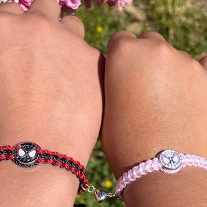 May include: Two matching friendship bracelets with a red and black braided cord and a pink and white braided cord. Each bracelet has a silver charm with a cartoon image of Spider-Man's mask.