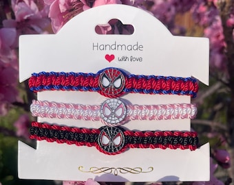 May include: Three handmade friendship bracelets with red, black and pink woven cords. Each bracelet features a silver-toned Spider-Man charm with a red and blue design. The card reads "Handmade with love".