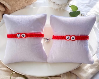 May include: Two red string bracelets with white beads that have the letters "A" and "J" on them. The bracelets are connected by a silver charm in the shape of a heart.
