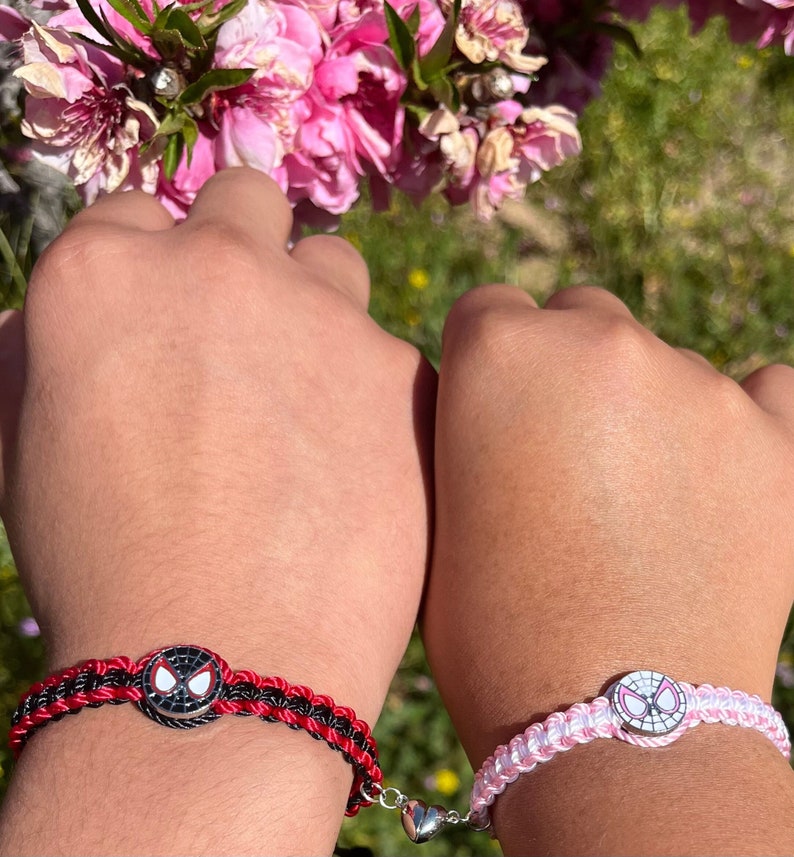May include: Two matching friendship bracelets with a red and black braided cord and a pink and white braided cord. Each bracelet has a silver charm with a cartoon image of Spider-Man's mask.