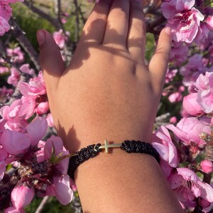 May include: Black braided bracelet with a gold-colored cross charm. The bracelet is worn on a wrist, surrounded by pink blossoms. The cross is centered on the bracelet.