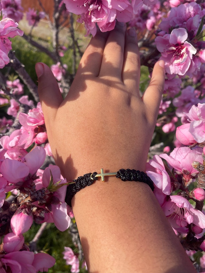 May include: Black braided bracelet with a gold-colored cross charm. The bracelet is worn on a wrist, surrounded by pink blossoms. The cross is centered on the bracelet.