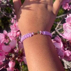 May include: A braided lavender bracelet with a gold cross charm. The bracelet is made of woven cord and features a small, gold-colored cross in the center. The bracelet is shown on a person's wrist, surrounded by pink flowers.