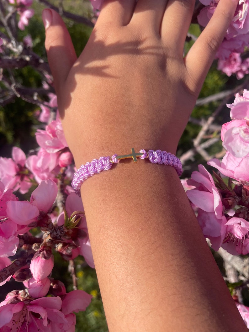 May include: A braided lavender bracelet with a gold cross charm. The bracelet is made of woven cord and features a small, gold-colored cross in the center. The bracelet is shown on a person's wrist, surrounded by pink flowers.