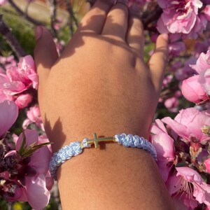 May include: A light blue macrame bracelet with a gold cross charm. The bracelet is displayed against a backdrop of pink blossoms, creating a soft and delicate aesthetic. The bracelet is a handmade accessory.