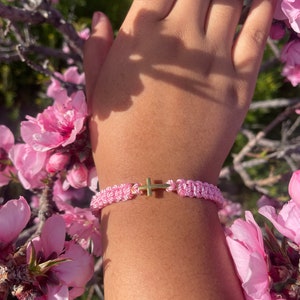 May include: Pink and white braided bracelet with a gold cross charm. The bracelet is displayed against a backdrop of pink blossoms and a person's arm.