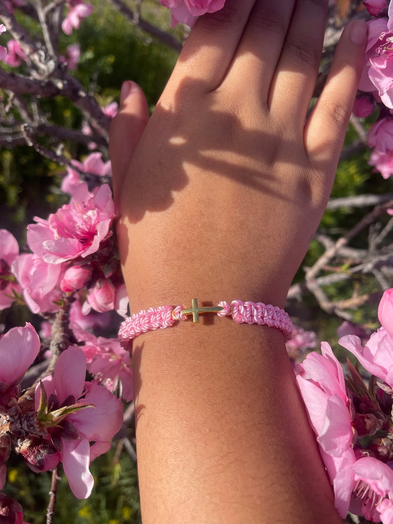 May include: Pink and white braided bracelet with a gold cross charm. The bracelet is displayed against a backdrop of pink blossoms and a person's arm.