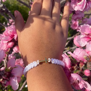 May include: White braided bracelet with a gold cross charm. The bracelet is made of woven white cord and features a small, gold-colored cross in the center. The bracelet is shown on a wrist, surrounded by pink flowers.