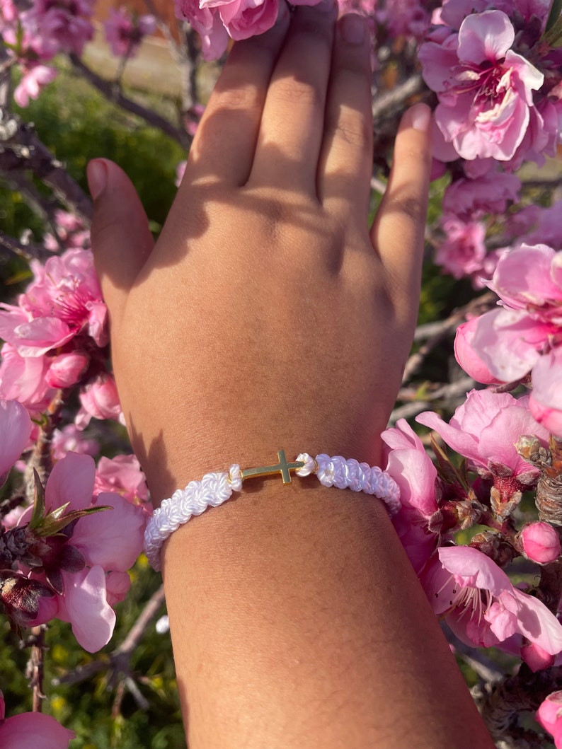 May include: White braided bracelet with a gold cross charm. The bracelet is made of woven white cord and features a small, gold-colored cross in the center. The bracelet is shown on a wrist, surrounded by pink flowers.