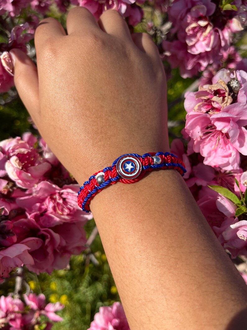 Puede incluir: Pulsera trenzada roja, blanca y azul con un dije de escudo de Capit&aacute;n Am&eacute;rica plateado.