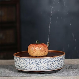 May include: A ceramic fountain with a pumpkin-shaped centerpiece. The pumpkin is orange and brown, with a green stem. It sits on a brown tray, which is set on a decorative base with a blue and white cloud pattern. Water sprays upwards from the pumpkin.