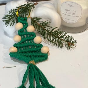 May include: Green macrame Christmas tree ornament with wooden beads and a gold tassel. The ornament is hanging from a gold key ring.