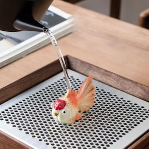 May include: A ceramic goldfish ornament with orange fins and a red head, positioned on a metal tray with a honeycomb design. Water is being poured from a black spout. The tray is set within a wooden frame.