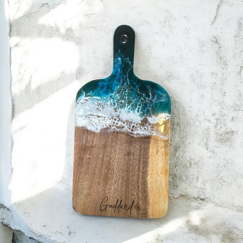 May include: A wooden serving board with a handle, featuring a decorative resin inlay at the top that mimics ocean waves in shades of teal, blue, and white. The natural wood grain is visible at the bottom, with the name Goddard printed in black.