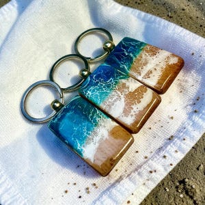 May include: Three rectangular keyrings with a beach scene design. The top half of each keyring features blue and white resin, resembling ocean waves. The bottom half is light brown wood, mimicking sand. Each keyring has a silver ring.