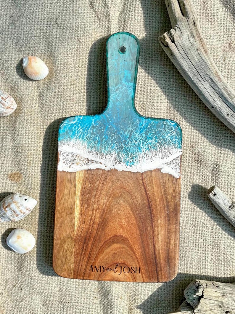 May include: A rectangular wooden serving board with a handle, featuring a blue and white resin ocean wave design at the top. The natural wood grain is visible on the lower portion, which is engraved with the text AMY and JOSH.