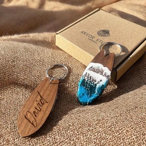 Puede incluir: Dos llaveros de madera con forma de tabla de surf. Un llavero tiene grabado el nombre "David". El otro tiene un diseño de olas oceánicas azules y blancas. Una caja de cartón con "ARVOR STUDIO CORNWALL" está en el fondo.