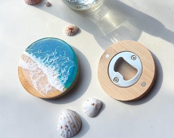 May include: Two round wooden coasters with ocean wave designs, one with a bottle opener. The coasters feature a beach scene with blue, white, and tan resin. Several seashells are scattered around the coasters.