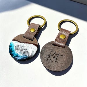 Könnte beinhalten: Zwei runde Holzschlüsselanhänger mit Messing-Schlüsselringen. Ein Schlüsselanhänger hat ein blau-weißes Harzdesign, das an Meereswellen erinnert. Der andere Schlüsselanhänger hat den Namen "Kat" auf der Holzoberfläche eingraviert. Beide Schlüsselanhänger haben braune Lederriemen.