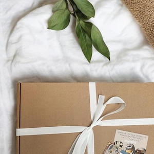 May include: A rectangular brown cardboard gift box tied with a white ribbon. The box features a small logo in the bottom left corner and two cards on the right, one labeled CARE GUIDE and the other reading UNIQUE, OCEAN-INSPIRED HANDCRAFTED GIFTS.