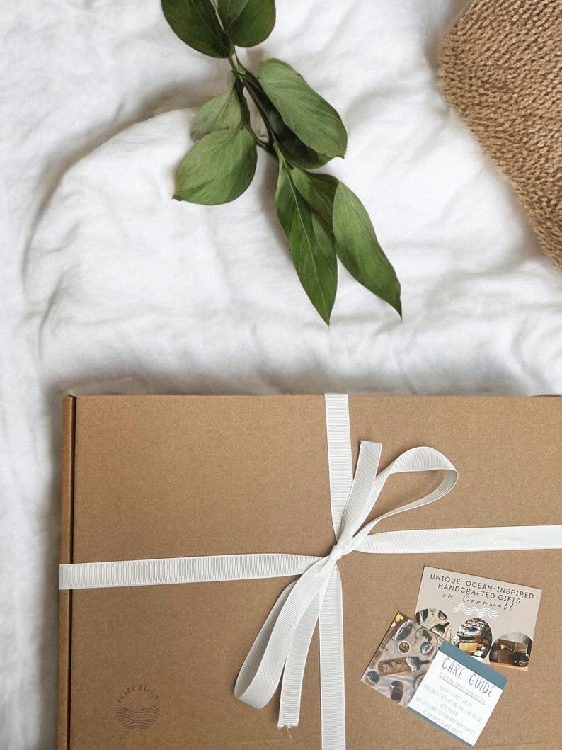 May include: A rectangular brown cardboard gift box tied with a white ribbon. The box features a small logo in the bottom left corner and two cards on the right, one labeled CARE GUIDE and the other reading UNIQUE, OCEAN-INSPIRED HANDCRAFTED GIFTS.