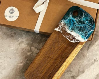 May include: Wooden serving board with a resin ocean design. The board features a natural wood grain base and a top section with blue, white, and brown resin, mimicking ocean waves. A brown cardboard box with a white ribbon and the "Arvor Studio" logo is in the background.