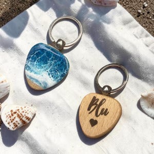 May include: Two keyrings with silver hoops. One has a blue and white ocean wave design over a wooden base. The other has the word "Blu" and a heart etched into a wooden heart shape. Seashells are scattered around.