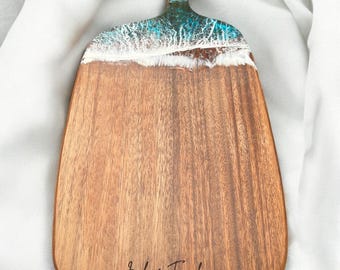 May include: Wooden serving board with a handle, featuring a unique ocean-inspired design. The top section has a blue and white resin wave pattern, transitioning to a natural wood grain. The board is engraved with "Jack & Frankie - 17.08.2019".