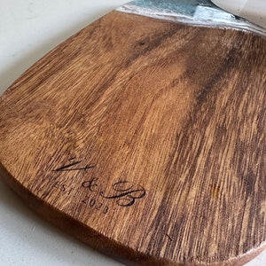 May include: A wooden serving board with a natural brown wood grain finish and a handle. The board features a decorative teal and white resin accent near the handle and is engraved with the text V & B EST. 2023 on the lower surface.