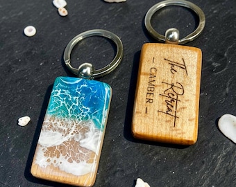 Könnte beinhalten: Zwei rechteckige Schlüsselanhänger. Einer zeigt eine Strandszene mit blauem, weißem und beigem Harz. Der andere ist aus Holz mit dem eingravierten Text "The Retreat - Camber". Beide haben silberne Schlüsselringe.