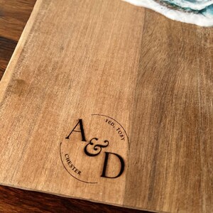 May include: A wooden serving board featuring a decorative resin ocean wave design in shades of blue and white at the top. The wood surface is engraved with a circular logo containing the letters A&D, along with the names Ted, Toby, and Chester.