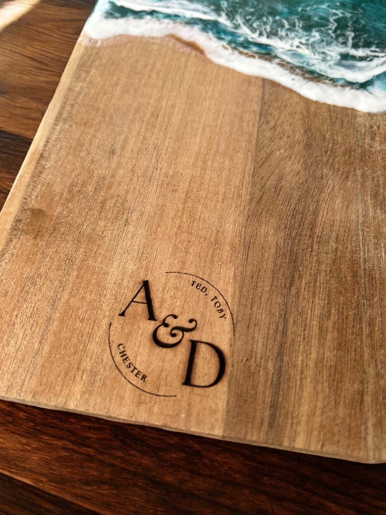 May include: A wooden serving board featuring a decorative resin ocean wave design in shades of blue and white at the top. The wood surface is engraved with a circular logo containing the letters A&D, along with the names Ted, Toby, and Chester.