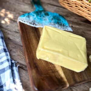May include: A rectangular block of pale yellow butter rests on a wooden serving board. The board features a dark brown wood grain and a decorative handle section finished with a glossy blue and white resin design resembling ocean waves.