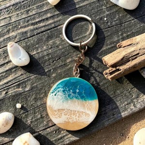 May include: A circular keychain with a beach-themed design. The keychain has a blue and white ocean design over a tan sand-coloured base. It is attached to a silver-coloured keyring and chain.