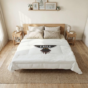 Puede incluir: Edredón blanco con un gran diseño de águila y la palabra "EAGLE" en negro. El águila tiene plumas marrones y negras. La cama tiene almohadas beige y una cabecera de madera. Dos mesitas de noche de madera con lámparas a ambos lados de la cama.