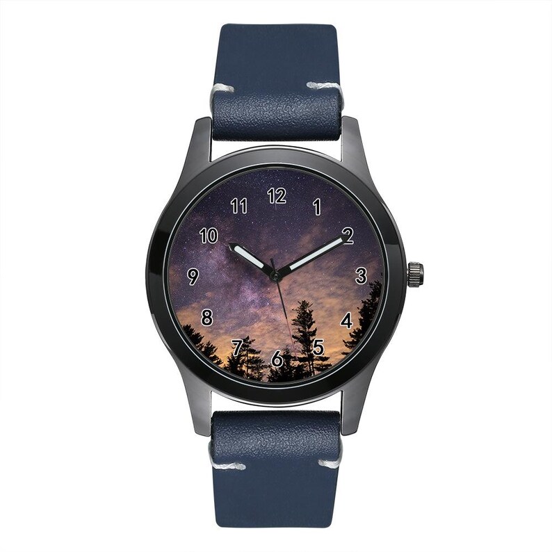 Pu&ograve; includere: Un orologio da polso con cinturino in pelle blu scuro e cassa in metallo nero. Il quadrante presenta un'immagine stampata di un cielo notturno con una galassia stellata, sagome di pini sul fondo e numeri arabi bianchi da 1 a 12.