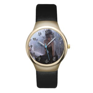 May include: A wristwatch with a gold-tone round case and a black leather strap. The watch face features a graphic illustration of a person in a black dress sitting in a rainy setting, with black Arabic numerals and silver hour, minute, and second hands.