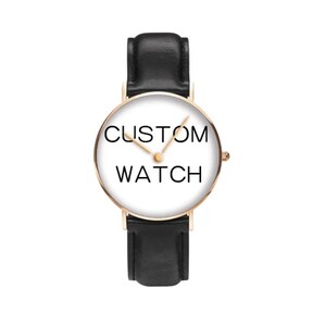Puede incluir: Un reloj de pulsera con caja redonda en oro rosa y esfera blanca con el texto negro CUSTOM WATCH. El reloj tiene manecillas finas en tono dorado y una correa de cuero negro liso con un cierre de hebilla negro a juego en la parte superior e inferior.