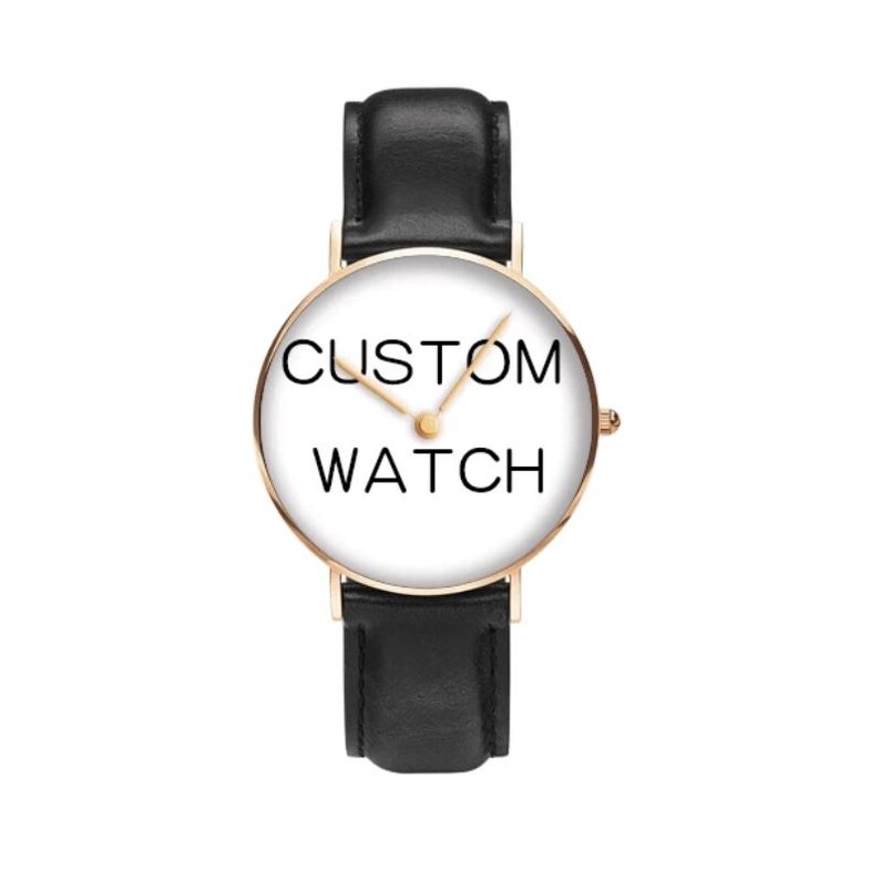 Puede incluir: Un reloj de pulsera con caja redonda en oro rosa y esfera blanca con el texto negro CUSTOM WATCH. El reloj tiene manecillas finas en tono dorado y una correa de cuero negro liso con un cierre de hebilla negro a juego en la parte superior e inferior.