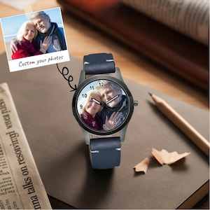 Pu&ograve; includere: Un orologio da polso con cinturino in pelle blu scuro e cassa nera. Il quadrante presenta una fotografia personalizzata di una coppia, con numeri neri da 1 a 12 e lancette bianche. Un piccolo riquadro mostra la fotografia originale.