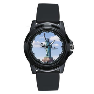 May include: A black wristwatch featuring a matte black case and a smooth black silicone strap. The watch face displays a photographic image of the Statue of Liberty against a blue sky with white clouds, surrounded by a black bezel with white numerals.
