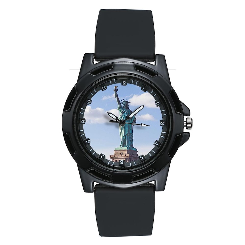 May include: A black wristwatch featuring a matte black case and a smooth black silicone strap. The watch face displays a photographic image of the Statue of Liberty against a blue sky with white clouds, surrounded by a black bezel with white numerals.