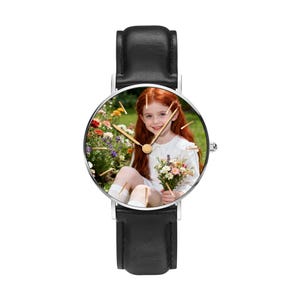 Puede incluir: Un reloj de pulsera con correa de cuero negro y caja circular plateada. La esfera del reloj presenta una impresi&oacute;n fotogr&aacute;fica personalizada de una persona joven con cabello largo y rojo, vistiendo una blusa blanca y sosteniendo flores.