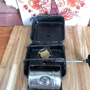 May include: A vintage camping stove with a black metal case, a fuel tank and a burner. The stove is open and ready for use.